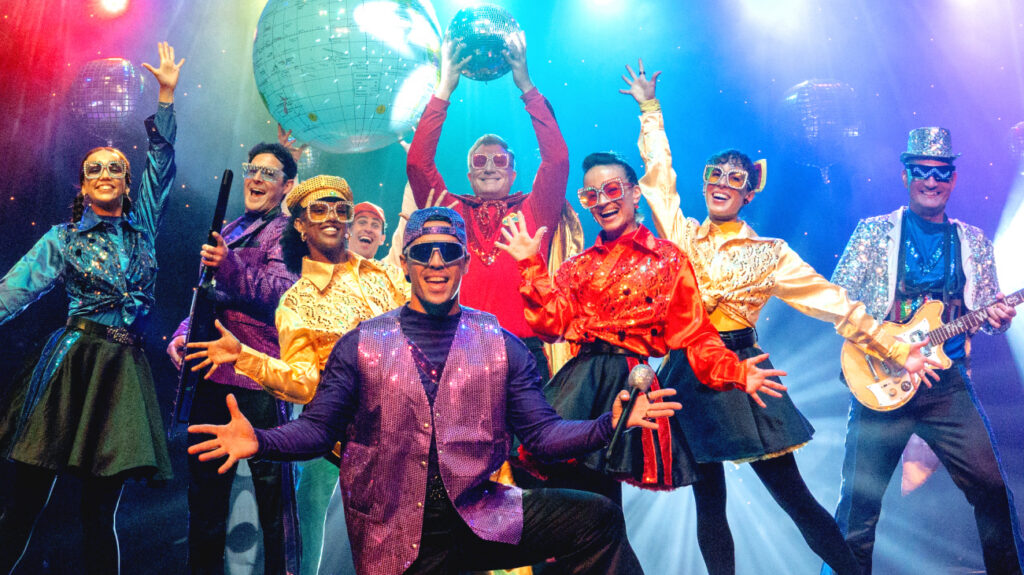 The Wiggles in colorful costumes on stage, entertaining an audience with music and dance.